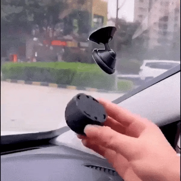 Hand holding a black car air freshener with a car's interior and windshield in the background